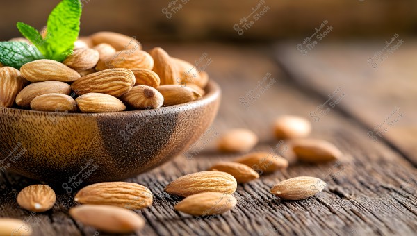 Image of almond seeds in a wooden bowl on a wooden surface. The bowl is filled with peeled almonds of a light brown color, with some almond seeds scattered on the table beside the bowl, and a green mint leaf decorates the top of the almonds in the bowl.