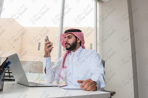 رجل سعودي يرتدي الزي السعودي التقليدي يعمل في مكتب بواجهة زجاجية ويستخدم قلم رصاص لتدوين الملاحظات