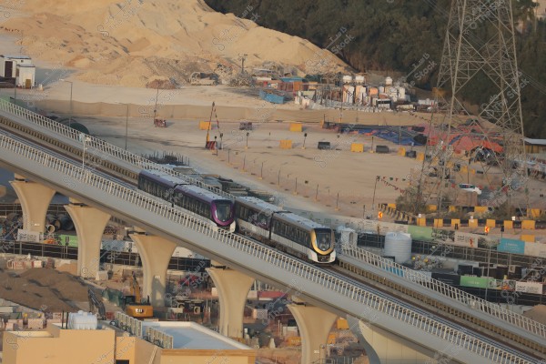 The image shows two trains traveling on an elevated bridge over a construction site with sandy terrain in the background. The site includes construction equipment such as excavators and concrete slabs. Behind the bridge, there is a desert landscape, trees, cables, and power towers.