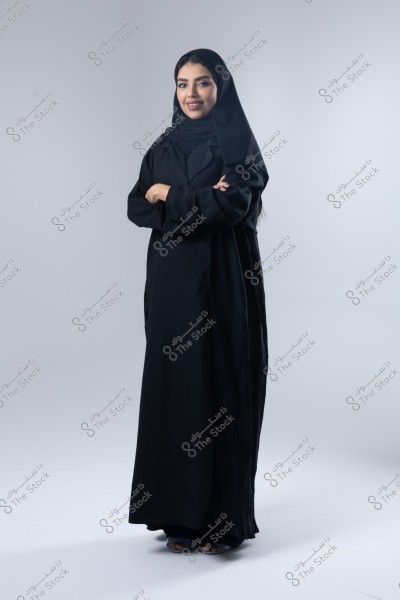 A portrait of a woman wearing a black abaya and hijab, standing with a smile and folded arms. The white background highlights the details of the traditional clothing.