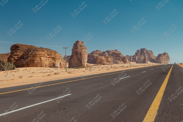 A scenic view of a paved road stretching along a desert in a mountainous area. Distinctive rock formations stand out in the background under a clear blue sky. There are also some power poles near the road, with sparse desert shrubs scattered on the sand.