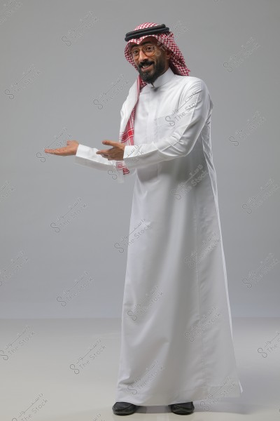 ** صورة لرجل واقف يرتدي ثوبًا أبيض وشماغًا أحمر وأبيض وعقالًا. الرجل يبتسم ويرفع يديه وكأنه يشير إلى شيء على يمينه. الخلفية رمادية بسيطة.\r\n\r\n**