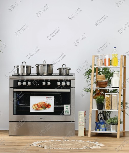 An image of a modern kitchen featuring a silver stove with three stainless steel cooking pots of varying sizes on top. Next to the stove is a wooden shelf holding preserved food in glass containers, cooking oil, wooden bowls, a blue kettle, and decorative plants. The background is white, with a wooden floor and a round wicker rug in front of the stove.