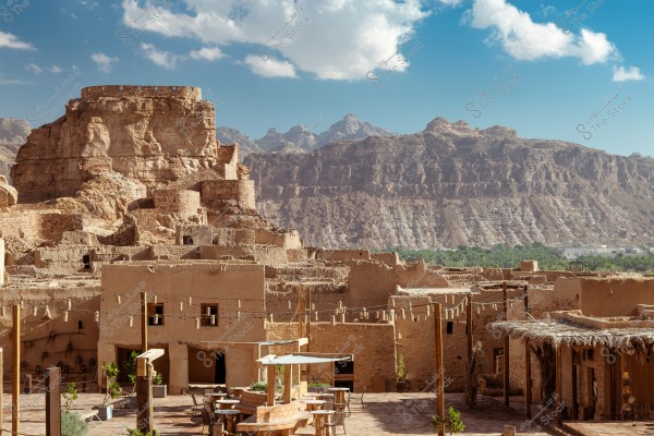 View of an ancient desert town with traditional mud buildings surrounded by hills and rocky formations. The sky is blue with scattered white clouds. The greenery in the background adds a natural contrast to the desert beauty.