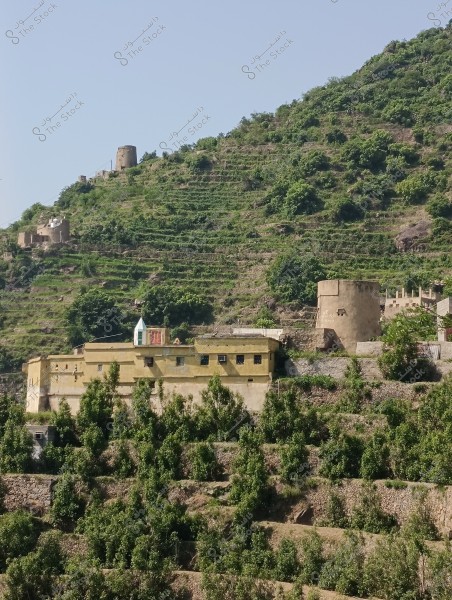 An image of a mountain village showing stone and mud houses situated on a slope planted with green trees and shrubs. The houses have a traditional design and are surrounded by terraced agricultural fields. In the background, an old stone tower can be seen atop the hill.