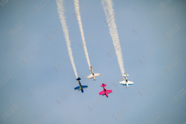 Four colorful airplanes flying in formation together in the clear sky, leaving trails of white smoke behind them. The airplanes are of different colors: blue, pink, white, and orange.