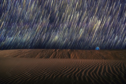 A shot of a tent in the middle of the desert whose sky is decorated with stars and meteors in a circular manner over the sand dunes in one of the Saudi deserts.