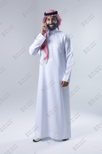 A portrait of a man wearing a white thobe and a red checkered headscarf (ghutra) with a black agal, standing and smiling while talking on the phone. The attire suggests he is likely of Saudi origin. The background is plain white.
