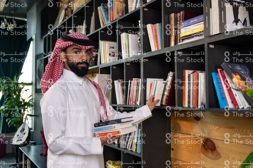 رجل سعودي يرتدي الزي السعودي التقليدي يقف في المكتبة بينما يحمل الكتب ويختار أحدهم