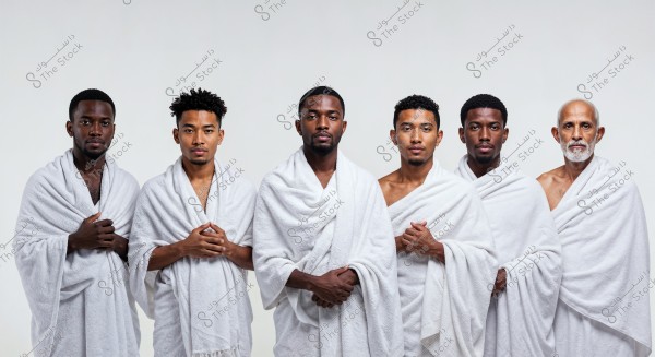 Six men stand side by side in a row, wearing white Ihram garments. They all face the camera against a bright white background, suggesting they are preparing for Hajj or Umrah. Their appearance is diverse, encompassing different ages.