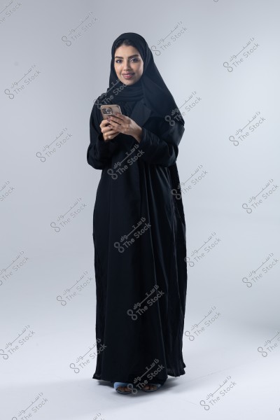 A portrait of a woman wearing a black abaya and hijab, standing in full-length against a neutral background. She is holding a mobile phone in her hands and smiling at the camera.