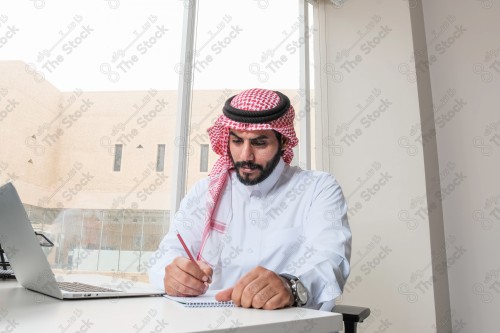 رجل سعودي يرتدي الزي السعودي التقليدي يعمل في مكتب بواجهة زجاجية ويستخدم قلم رصاص لتدوين الملاحظات