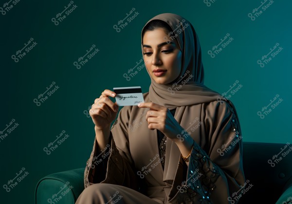 Image of a woman sitting on a green couch, wearing a brown abaya with embroidery. The woman is veiled and looking at a plastic card with a signature. The background is dark green.