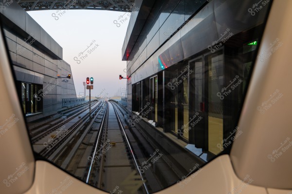 View from inside a train showing the railway tracks extending between a building with dark glass and a modern metal structure. Railway traffic signals in green and red are also visible. The sky is a light background color in the distance.