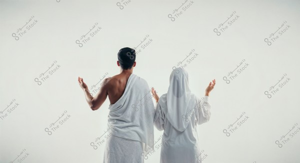 Two individuals stand in prayer, one man wearing a white Ihram and a woman in a white hijab and clothing. The background is plain white.