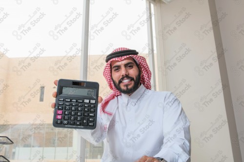 رجل سعودي يرتدي الزي السعودي التقليدي يعمل في مكتب بواجهة زجاجية ويستخدم آلة الحاسبة أثناء العمل