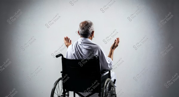 A photo of an elderly man sitting in a wheelchair wearing a white Ihram garment, with his hands raised in a praying posture, against a plain white background. The image captures a moment of devotion and spirituality.