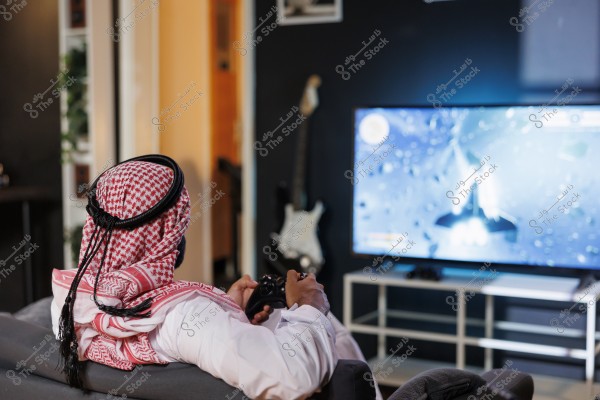 A man wearing traditional clothing and a red ghutra is sitting on a couch holding a video game controller, focused on a large TV screen displaying a space-themed video game. In the background, there are dark walls with an electric guitar hanging and framed pictures.