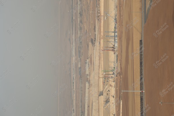 An image of a large construction site in a desert area. Construction equipment such as cranes are visible in the center of the image, with signboards reading \"The Largest Modern Downtown.\" A light haze fills the horizon in the background, indicating the arid atmosphere of the location.