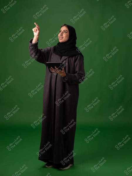 An image of a woman wearing a black abaya and hijab, standing in front of a green background. She is holding a tablet in one hand and pointing upward with the other as if indicating something. She is wearing black athletic shoes.