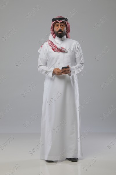 رجل يرتدي ثوبًا أبيض وغترة حمراء وبيضاء مع عقال أسود, يمسك بهاتف محمول. يبدو أنه يقف في استوديو ذو خلفية رمادية بسيطة.