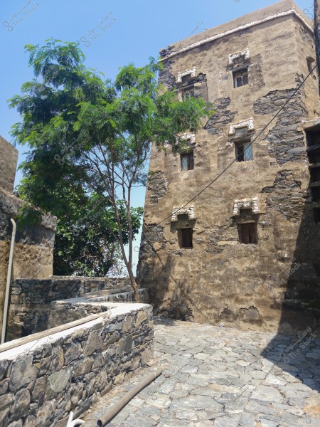 The image depicts an old multi-story mud building with small windows, highlighting a traditional design with exposed stones. Next to the building, a green tree adds a natural touch to the scene. The ground is paved with natural stones, and some pipes are visible on the ground. The sky is clear and blue in the background.