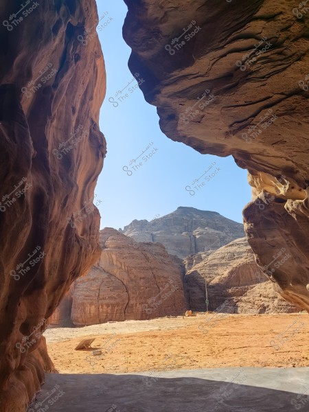 The image shows a natural landscape from inside a cave, featuring multi-colored rock walls with a view of a mountain in the distance under a clear sky. The ground is sandy and rocky, representing a desert landscape in a mountainous area.