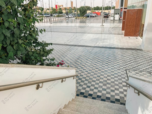 The image shows a stone staircase covered with multi-colored patterned tiles, with metal railings on both sides. In the foreground, there is a large green tree with dense leaves and red flowers. In the background, a wide plaza is visible, covered with square colored tiles, and a parking lot with several cars. Buildings can be seen in the horizon under clear skies.
