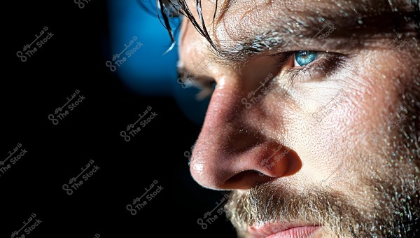 Close-up image of a man\'s face with a thick beard, focusing on his blue eye which looks intently to the right. The man\'s face appears sweaty under bright lighting, highlighting the texture of his skin. The dark background accentuates the facial features dramatically.