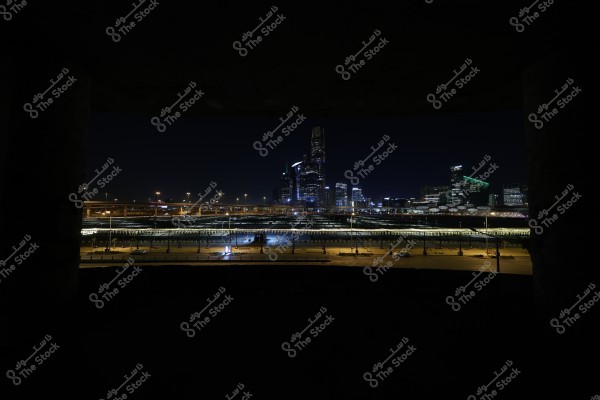 Nighttime view of a modern city featuring skyscrapers illuminated with colorful lights. In the foreground, there is an elevated, lit bridge with vehicles traveling beneath it. The sky is dark, emphasizing the bright lights of the buildings.