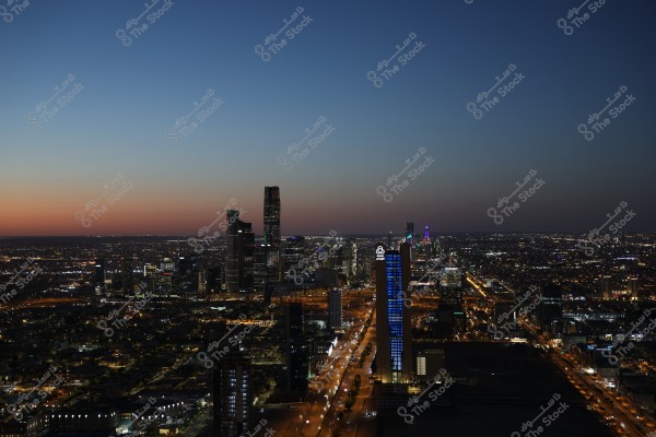An image of a modern city at dusk, featuring sparkling city lights and a skyline of tall buildings. A tall tower in the foreground is illuminated with blue lights, and the sun is setting on the horizon, adding shades of orange to the dark blue sky.