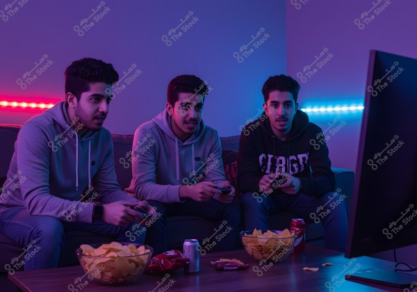 Three young men seated on a couch in a room illuminated by blue and red neon lighting, playing with video game controllers and focused on a TV screen in front of them. In front of them on the table are bowls filled with chips and cans of soda. The young men are dressed in casual athletic hoodies, suggesting a relaxed and sociable gathering.