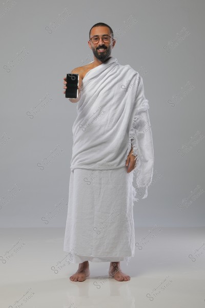 A portrait of a man wearing a white Hajj ihram, standing on a white floor. He is smiling and holding a smartphone in his right hand. He is wearing glasses and has a short beard. The background is light gray.