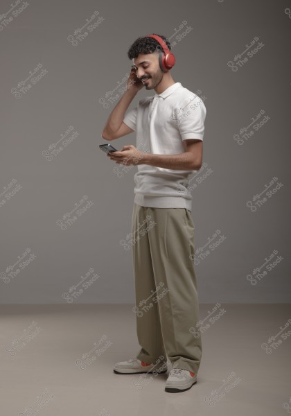 A man wearing a white t-shirt and beige trousers is smiling while looking at a mobile phone in his hand. He is wearing red headphones, appearing to enjoy music in a neutrally lit indoor space.