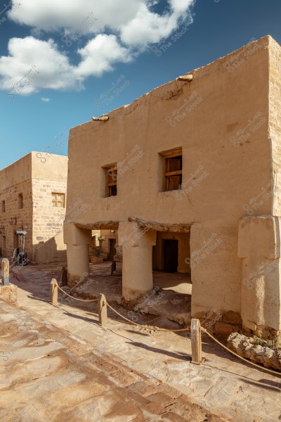 A traditional two-story mud building with small exterior columns. The building features wooden windows and large doors, designed in a traditional Arabic architectural style. The blue sky with a few white clouds is visible in the background, giving the image a sense of calm and tranquility.