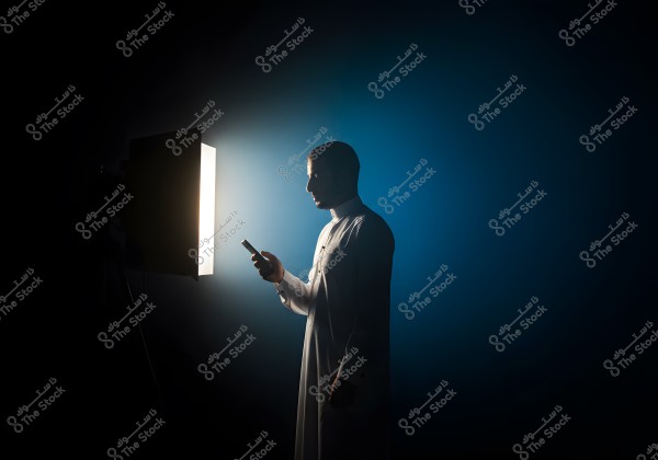 A portrait of a man wearing a traditional white thawb standing sideways while holding a smartphone. A strong light is focused on him from the left, creating a dramatic effect against a dark blue background. The intense play of light and shadow gives the image a cinematic atmosphere.