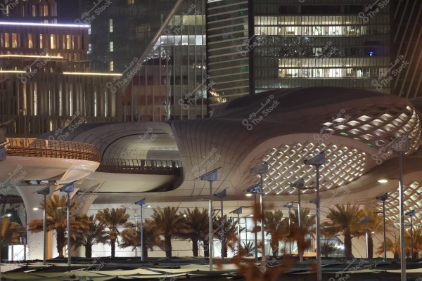 Nighttime image of a modern architectural building in a city. The building is illuminated with night lights and features geometric openings, giving it a unique appearance. Palm trees in front of the building add a natural touch to the urban setting.