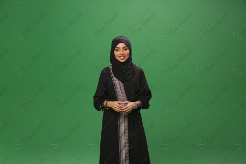 Saudi woman on green background wearing abaya and hijab, making movements with her hands, holding her hand while smiling, Eid celebrations