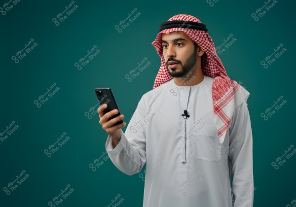 An image of a man wearing a traditional white thobe and a red and white shemagh. He is holding a smartphone and appears to be standing. The background is a solid green, emphasizing the person in traditional Gulf attire.