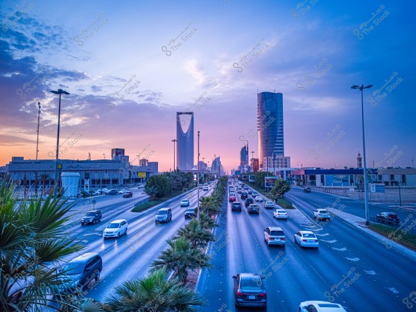 منظر لمدينة الرياض عند غروب الشمس, يظهر برج المملكة الأيقوني في وسط الصورة محاطًا بالمباني الحديثة الأخرى. الطريق السريع الرئيسي يمتلئ بالسيارات حيث تتوجه في كلا الاتجاهين, محاطًا بأشجار النخيل على الجانبين. السماء ملونة بألوان أرجوانية وزرقاء مما يضفي جمالية على المشهد الحضري.