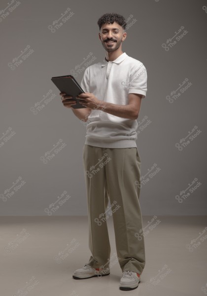 The image shows a young man standing and smiling with a tablet in his hands. He is wearing a white polo shirt and beige trousers. He appears to be in a studio with a neutral gray background. The young man is wearing white sneakers.
