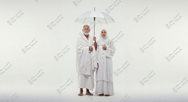 An image of a man and woman standing together in white Ihram clothing, representing the attire for Hajj. The man is wearing a white wrap and robe, while the woman is in a white abaya and headscarf. They are holding an open white umbrella, set against a white background.