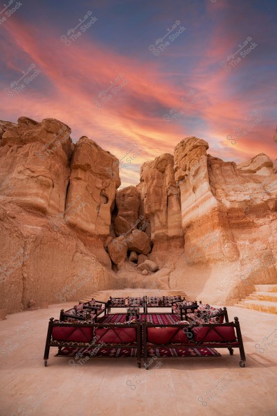 The image shows a stunning landscape in a desert area under a breathtaking sunset sky. Large, mountainous rock formations create a striking backdrop, sculpted by the wind into unique shapes. In the foreground, there is a traditional Arabic majlis, furnished with sofas covered in brightly colored patterned fabrics, arranged to welcome visitors amidst this enchanting natural scene.