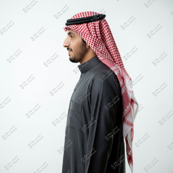 A side profile portrait of a man wearing a dark-colored thobe with a red and white checkered shemagh and a black agal. The man is set against a white background.