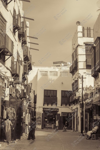 A black and white image of an old street showcasing traditional architectural style in the buildings. Wooden windows and wall decorations are visible. A few people are walking in the street, one wearing traditional attire. There\'s a sign with the text \"الشلي للعطارة\".
