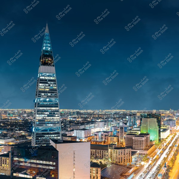 صورة ليلية لمدينة الرياض في المملكة العربية السعودية, تظهر فيها برج الفيصلية مضاءً بأنوار زرقاء وبيضاء. البرج هو مبنى طويل ذو تصميم هرمي مع قمة حادة, ويحيط به مبانٍ ومتاجر مضاءة بأضواء ليلية مبهرة. السماء داكنة وتبرز الأبراج والمعمار الفاخر للمدينة.