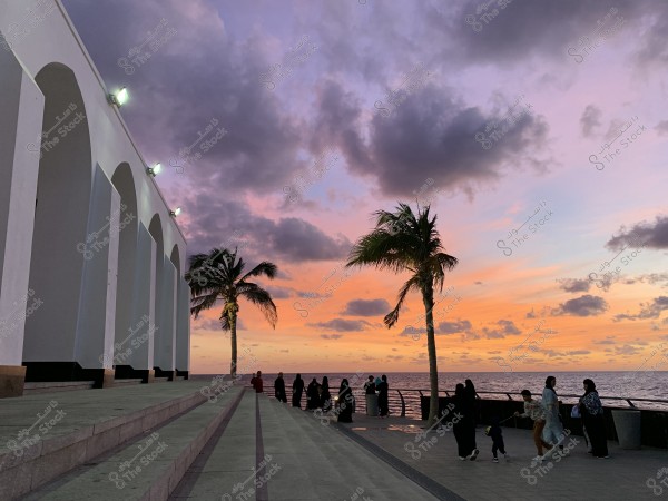 An image showing a sunset over a seaside promenade in Jeddah with an arched building lit on the left, and a palm tree on the right near the sea. A group of people are walking or sitting near the promenade\'s railing, and the horizon is adorned with vibrant orange and purple colors with scattered clouds.