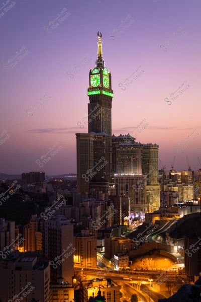 Image of the city skyline of Mecca at sunset, featuring the iconic Clock Tower with its illuminated green and gold facade. The building is surrounded by numerous residential and commercial buildings, with some lit roads visible in the foreground. The sky is a warm purple hue, reflecting the dazzling city lights.
