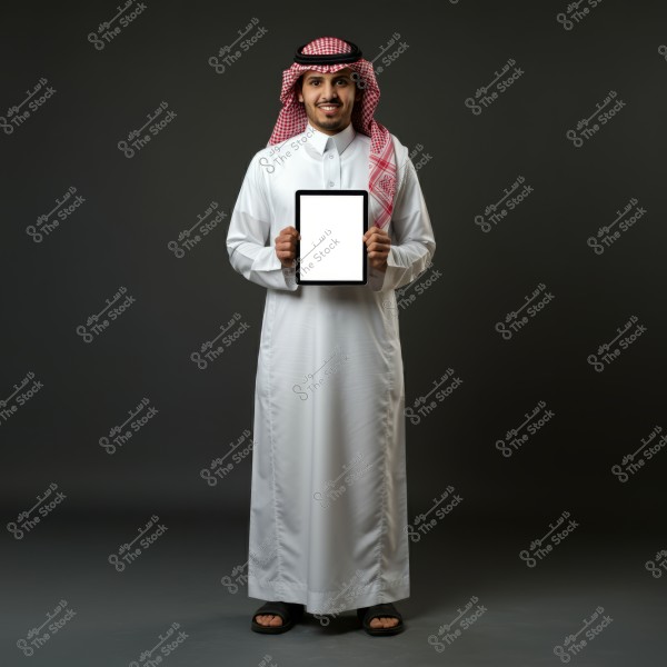 Image of a man standing against a dark background wearing a white thobe and a red checkered ghutra with an agal, indicating traditional Saudi attire. The man is holding a tablet with a blank white screen and appears to be smiling.