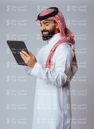 بورتريه صورة جانبية لرجل سعودي يرتدي الزي السعودي التقليدي يمسك بيده الآيباد و ينظر لشاشته، شاب مبتسم يستخدم الجهاز اللوحي